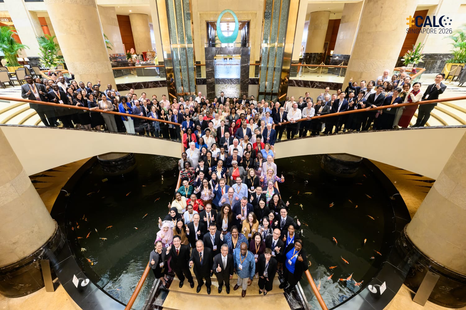 A large group of CALC Singapore 2026 conference delegates pose for a group photo in a grand hotel atrium, gathered around a koi pond and across two levels of balconies.